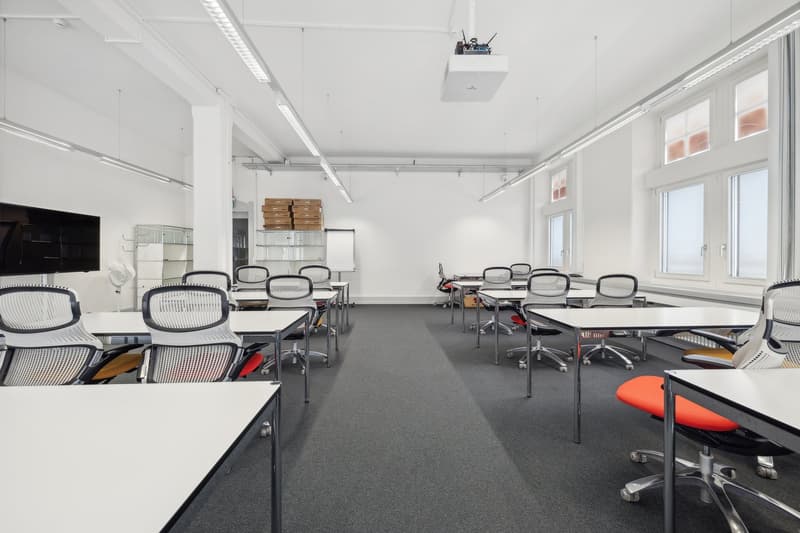 empty classroom, desks and chairs in rows, white walls, gray floor, large windows, whiteboard, TV mounted on wall, ceiling lights