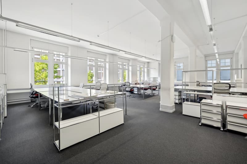 A modern office space featuring long tables with chairs, metal shelves, and large windows with natural light coming in. The flooring is carpeted, and the walls and ceiling are white. There are multiple office areas with tables and chairs visible.