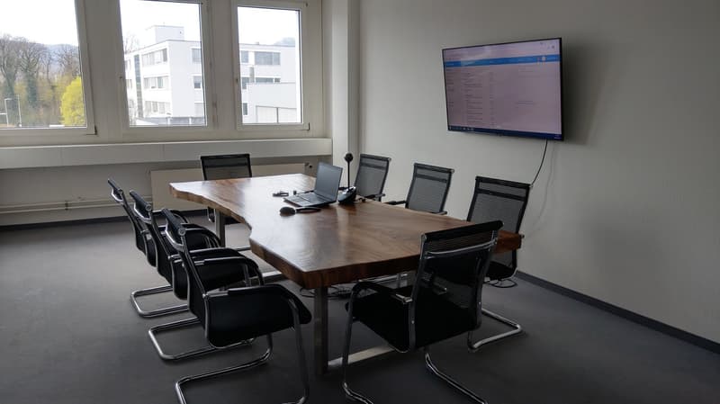 An empty conference room with large windows. It features a large wooden desk, modern black chairs, and a mounted screen on the wall. The room has gray flooring and a view of the outside from the windows.