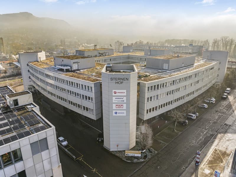 Aerial view of a large multistory office building with a green roof, multiple companies named on the side, surrounded by trees and parked cars