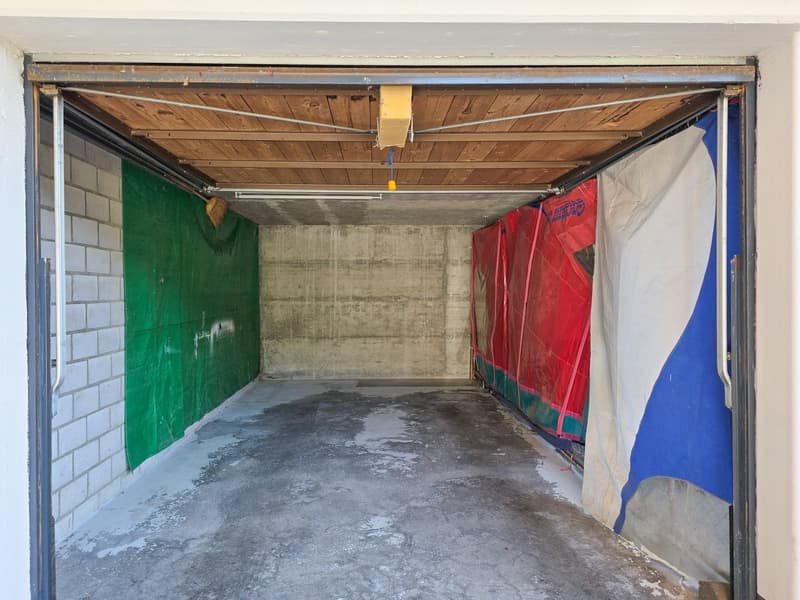 Concrete floor, open doorway, wooden ceiling, two walls, one covered with green tarpaulin, other with red and white fabric