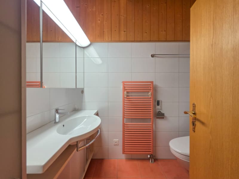 A white sink with a faucet, a white mirror above the sink, a toilet, and a towel rack against the wall. A bright light on the ceiling and wooden walls. Tiled floor.