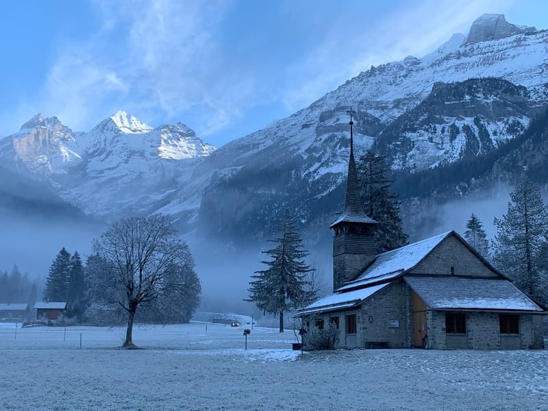 Small stone church with a steeple, surrounded by snow-covered mountains and trees