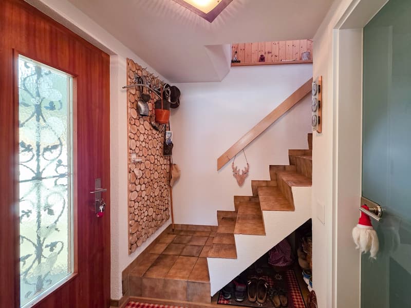 Entrance with wooden door, glass window, tiled floor, wooden stairs, wall with logs, storage under stairs, wooden handrail, wooden shelf, decorative items