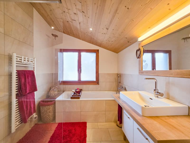 Bathroom with bathtub, sink, mirror, wooden ceiling, tiled floor, towel rack, red towels, red rug, wooden counter, white cabinets, wooden basket, and a window.