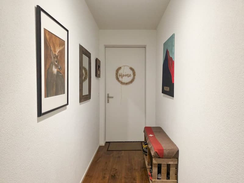 A hallway with white walls, wooden floor, a white door, a bench, and a shoe rack. There are three framed pictures on the wall, one of which has a wreath with the word 'Home' on it.