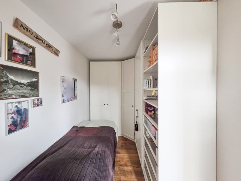 Small bedroom with bed, white walls, wooden floor, white wardrobes, shelves, and framed pictures