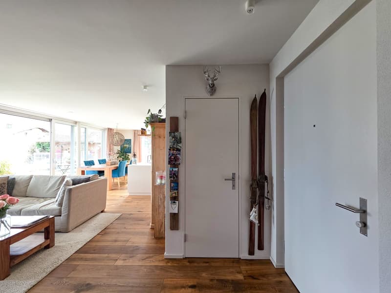 living room with wooden floor, white walls, couch, coffee table, rug, deer head, skis, photo frame, glass windows, dining table, chairs, plant