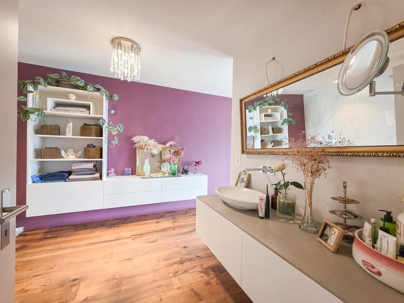 Bathroom with white cabinets, purple wall, wooden floor, round mirror, sink, shelves with various items, chandelier, and plants