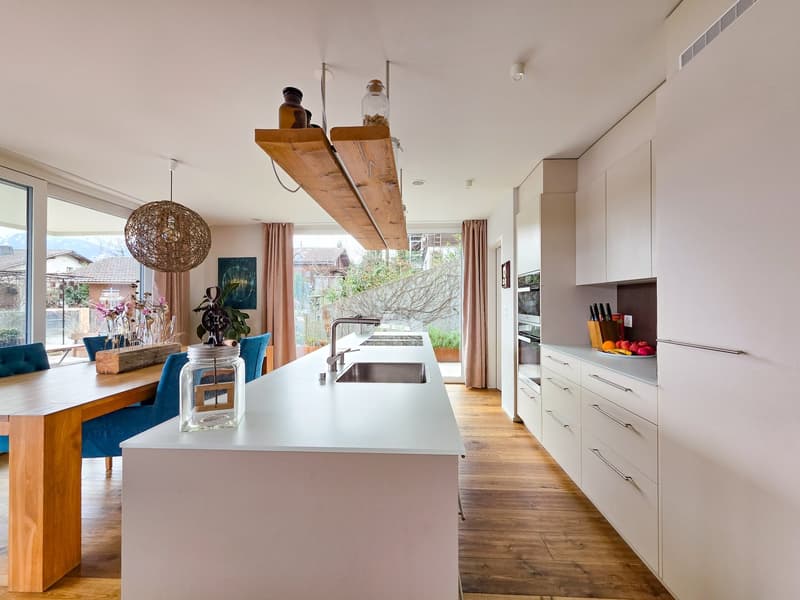 modern kitchen with wooden floor, white countertops, island sink, wooden dining table, blue chairs, large windows, wooden shelves, hanging lamp, plants, artwork