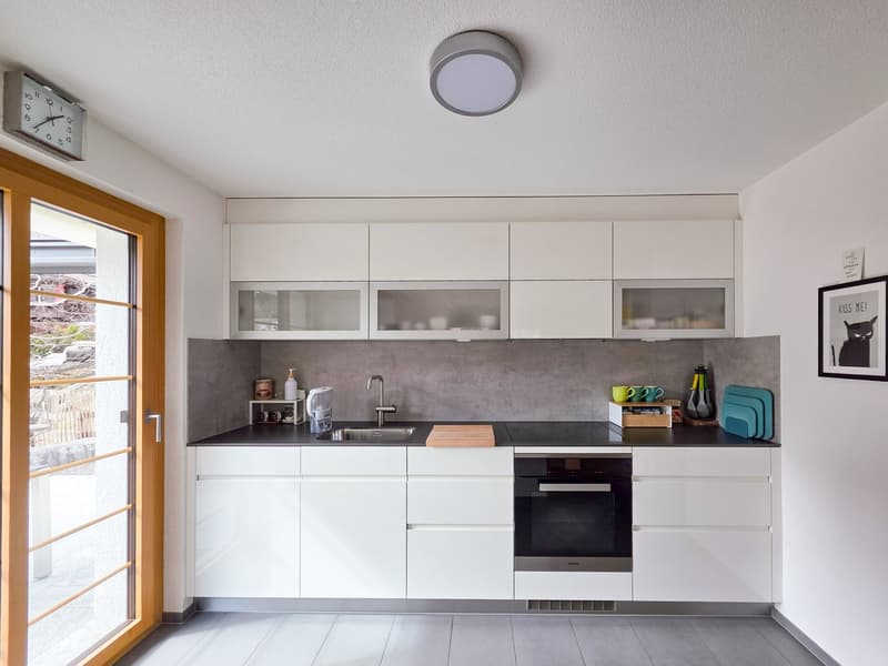 modern kitchen with white cabinets, black countertops, stainless steel sink, oven, and microwave, gray backsplash, ceiling light, and a glass door leading to a patio