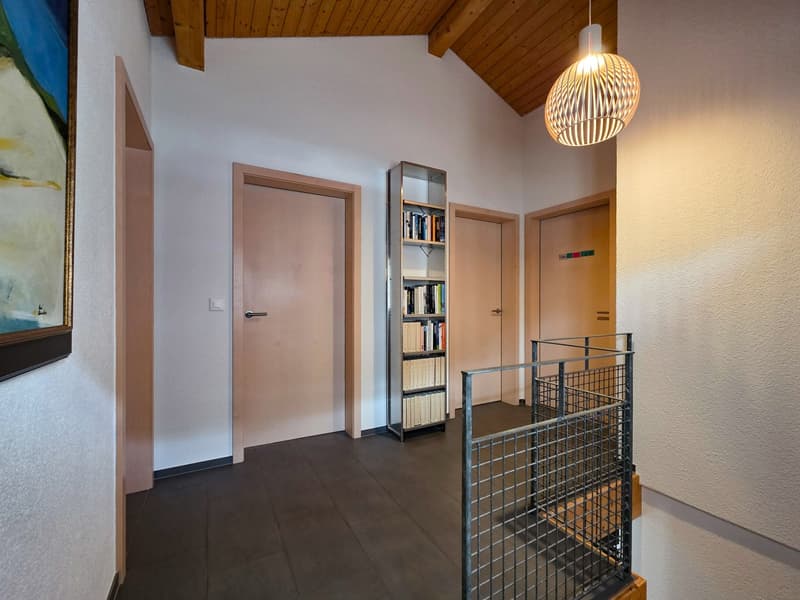 Empty hallway with wooden ceiling, white walls, tiled floor, metal railing, wooden doors, bookshelf, painting, light fixture