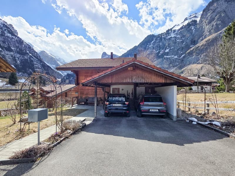 Wooden house, covered garage, two cars parked, mountains in the background