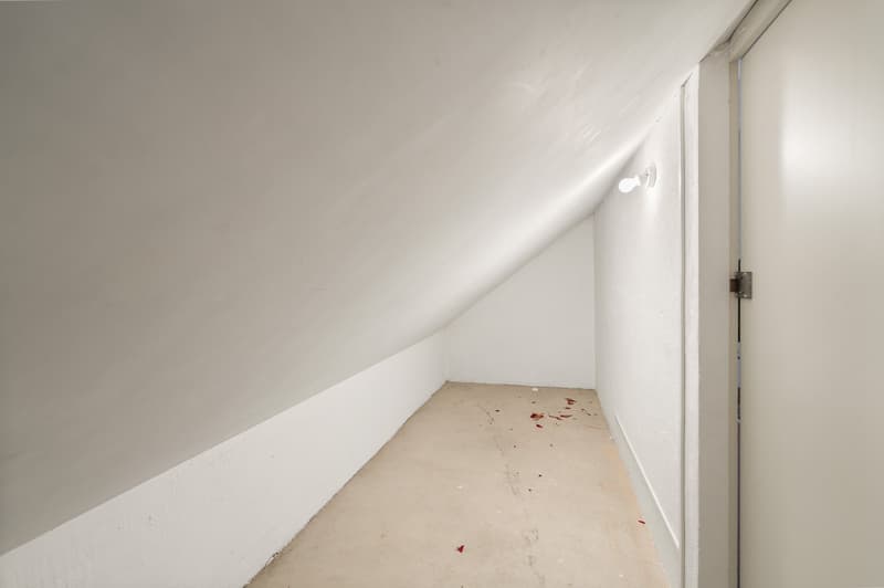 A sloped ceiling room with a white door, a ceiling lamp, and a floor with debris.