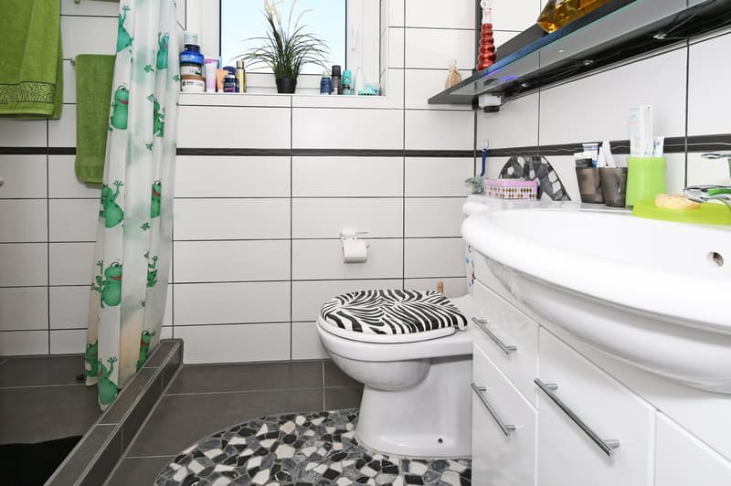 Bathroom with white walls and tiles, black countertop, shower curtain with frog design, white toilet, sink, and storage shelves, and black and white rug on the floor.