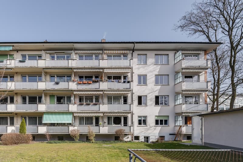 multi-story building, white exterior, multiple balconies, green awnings, fenced yard, trees