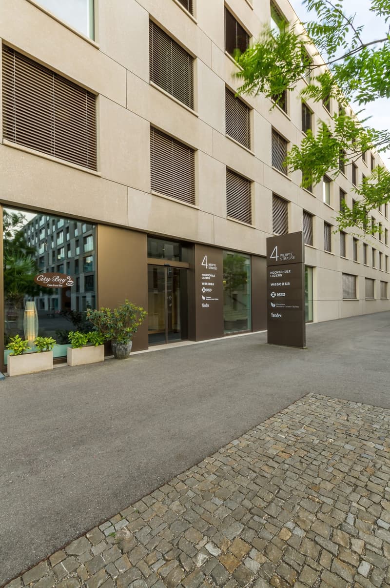 Modern building with glass walls and brown facades, several floors, entrance with automatic doors, plants, and pavement.