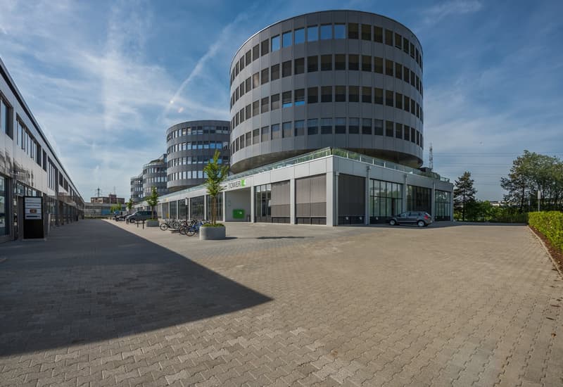 A modern, multi-story office building with a curved, glass facade. The building has a ground floor entrance and a plaza area with bicycle racks. There are several cars parked in the area in front of the building.
