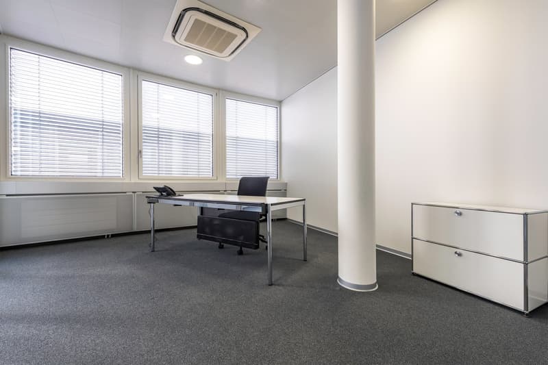 The image shows a modern office space with a large desk, a black office chair, and a filing cabinet. The room has large windows with blinds, providing natural light. The walls are painted in a light color, and the floor is covered with gray carpeting. The
