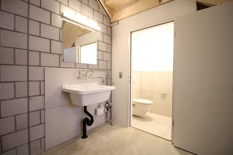 tiled walls, concrete floor, white sink with faucet, mirror, toilet, power outlet, exposed plumbing