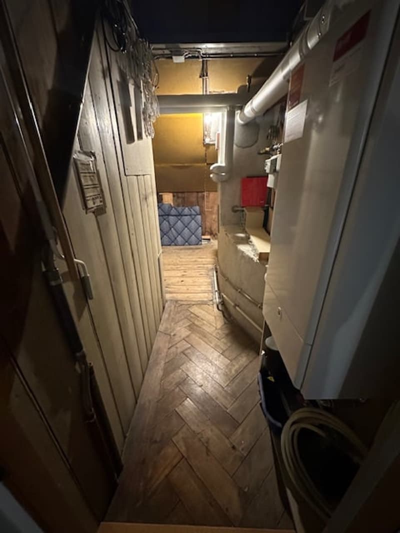 A narrow hallway with wooden flooring, a blue mattress, and a white pipe on the wall.