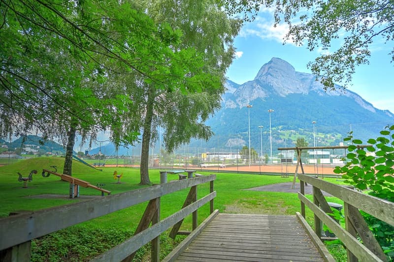The image shows a scenic outdoor park or recreation area with a wooden walkway, lush green grass, and trees. In the background, a large mountain peak is visible, indicating a picturesque natural setting. The area appears to be well-maintained and suitable