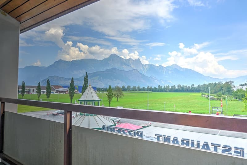 The image shows a balcony or terrace overlooking a scenic landscape. In the distance, there are snow-capped mountains and a grassy field with trees. The balcony has a wooden roof and railing, and there is a sign for a restaurant or establishment visible.