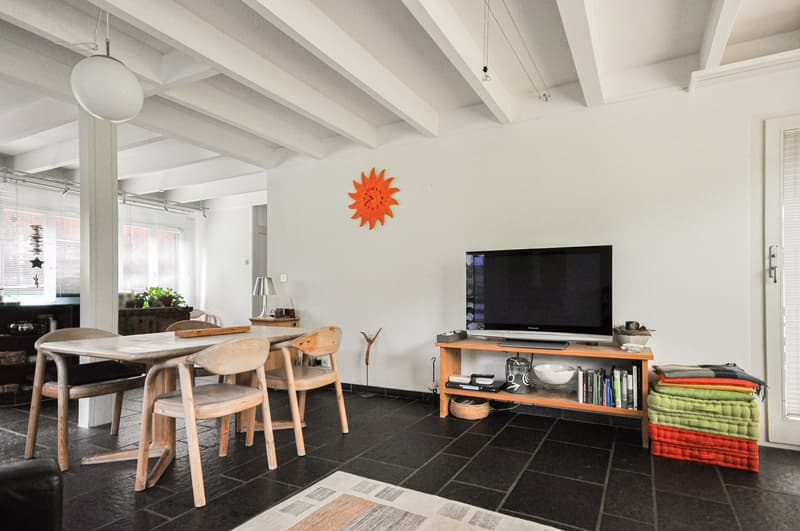 Dining and living room with dining table, chairs, TV and books on shelf, black tiled floor, hanging lamp and wall clock.