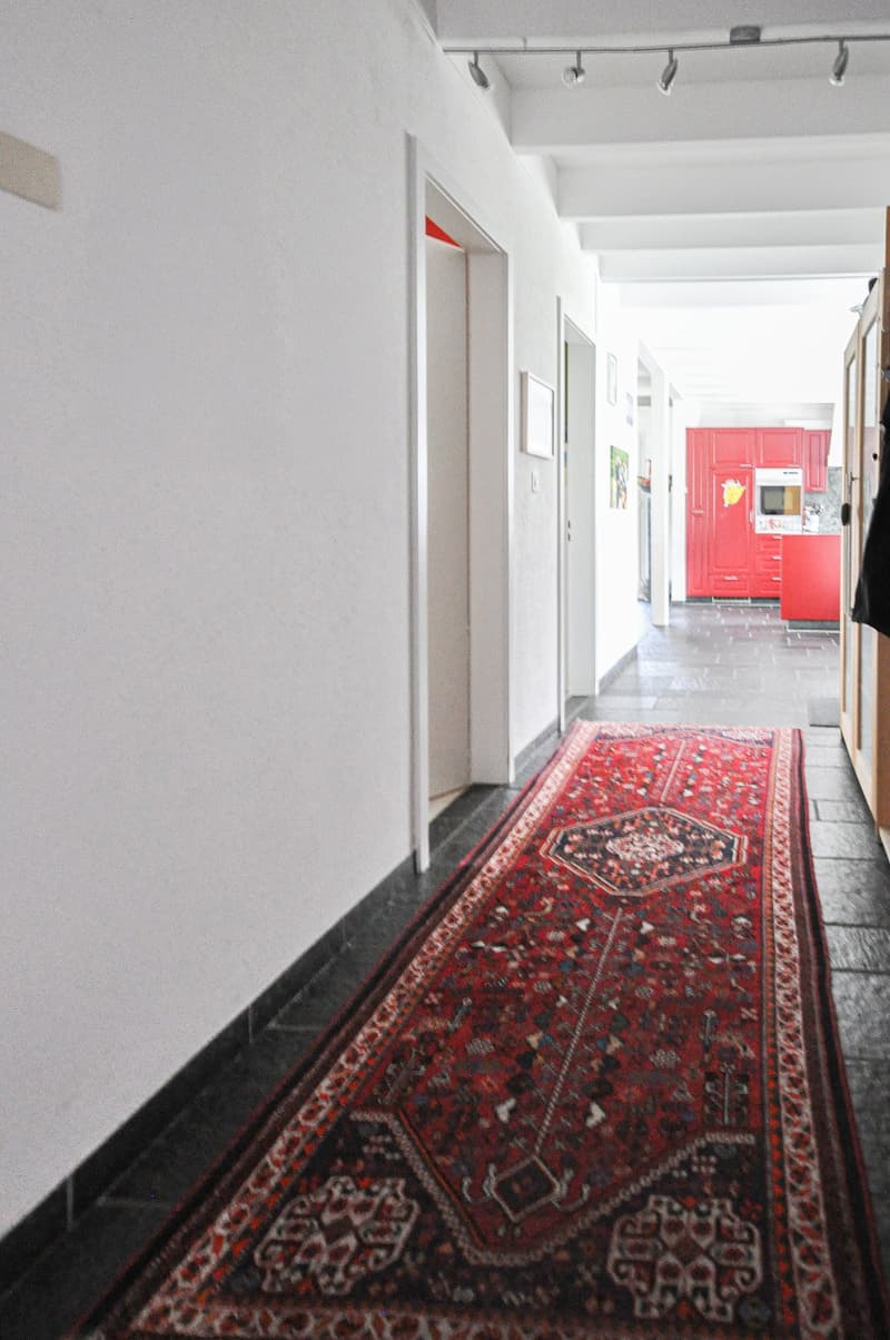 narrow hallway with white walls, tile flooring, red rug, open doorways, a red kitchen