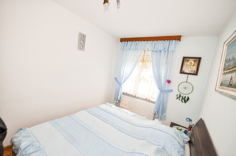 The image shows a bedroom with white walls, a wooden beam on the ceiling, and blue curtains with a floral pattern. There is a bed with a blue and white striped comforter, a nightstand, and a framed artwork on the wall. A dreamcatcher decoration is also vi