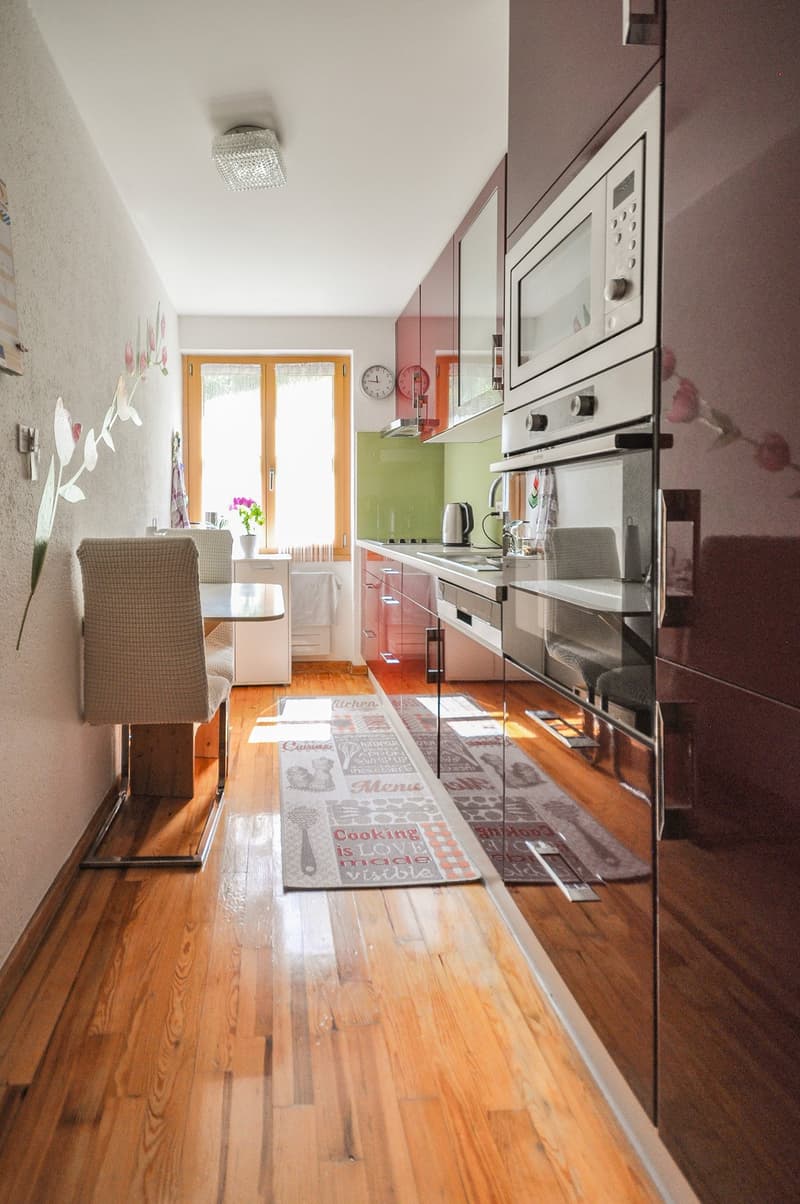 Spacious kitchen with hardwood floors, white cabinets, stainless steel appliances including a dishwasher, and a decorative kitchen rug on the floor.