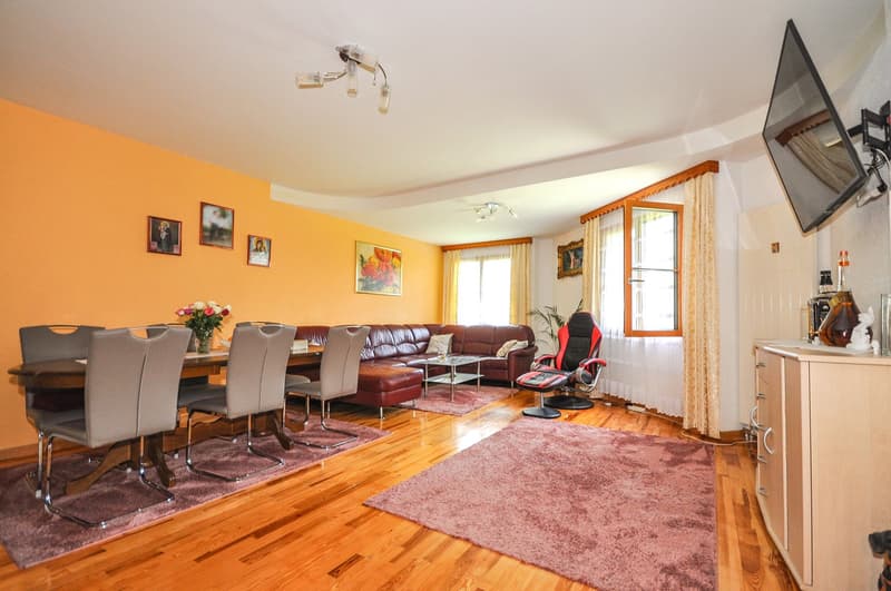 Spacious living room with yellow walls, hardwood floors, large windows, leather sofa and armchair, dining table with chairs, and a fireplace. The room is decorated with artwork and has a cozy, inviting atmosphere.