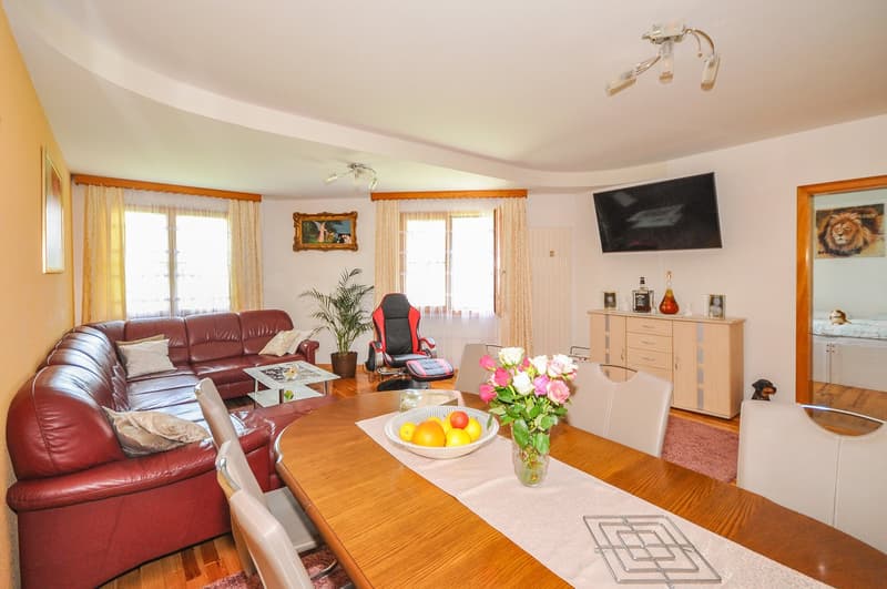 Spacious living room with large windows, red leather sofa, wooden dining table, TV, and decorative items such as a vase of flowers and a painting on the wall.
