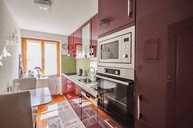 Modern kitchen with red cabinets, stainless steel appliances including a microwave and oven, hardwood floors, and a window providing natural light.