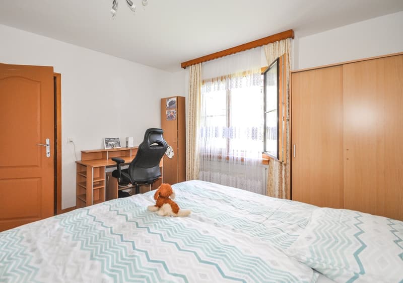 Spacious bedroom with large windows, wooden furniture including a desk and wardrobe, and a plush toy on the bed.
