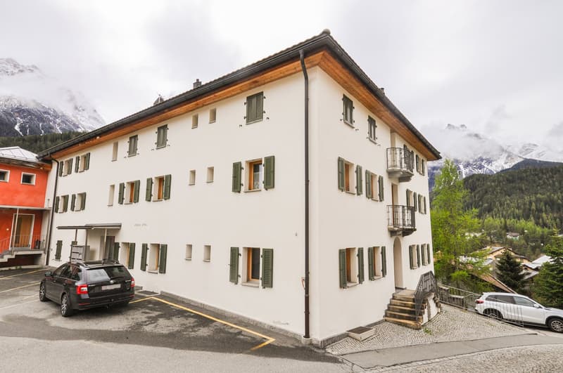 3-story apartment building with white exterior, green shutters, and a slanted roof. There is a parking area in front of the building with a few cars parked. The building is situated in a mountainous, forested area with snow-capped peaks visible in the bac