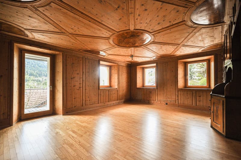 Large open room with wooden paneled walls and ceiling, hardwood floors, and large windows providing a scenic view. The room has a rustic, cabin-like aesthetic.