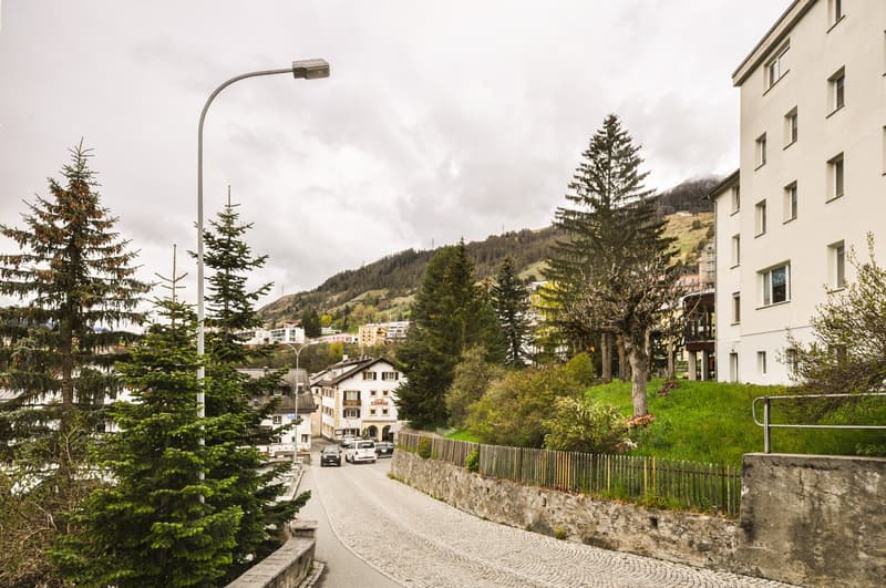 The image shows a residential neighborhood in a mountainous area. There are several multi-story apartment buildings, trees, and a winding road with parked cars. The buildings have balconies visible, and the surrounding landscape includes hills and forests