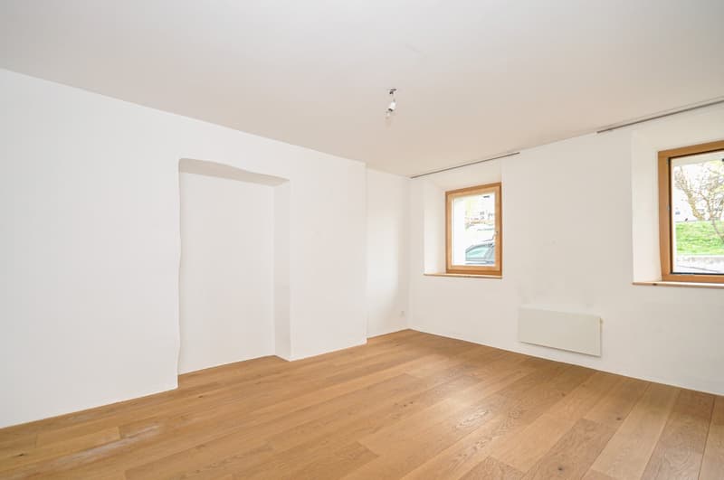This image shows an empty room with white walls and a hardwood floor. The room has two windows that provide natural light, and there is a heating unit visible on the wall. The room appears to be a living room or bedroom, with a simple and minimalist desig