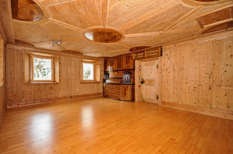 This image shows a large, open-concept room with a wooden interior. The room has a high, domed ceiling with intricate wooden paneling and a central light fixture. The walls are also made of wood, and there are two large windows that provide natural light.