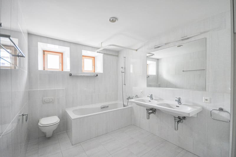 Spacious bathroom with white tiles, a bathtub, a double sink vanity, and large windows providing natural light.