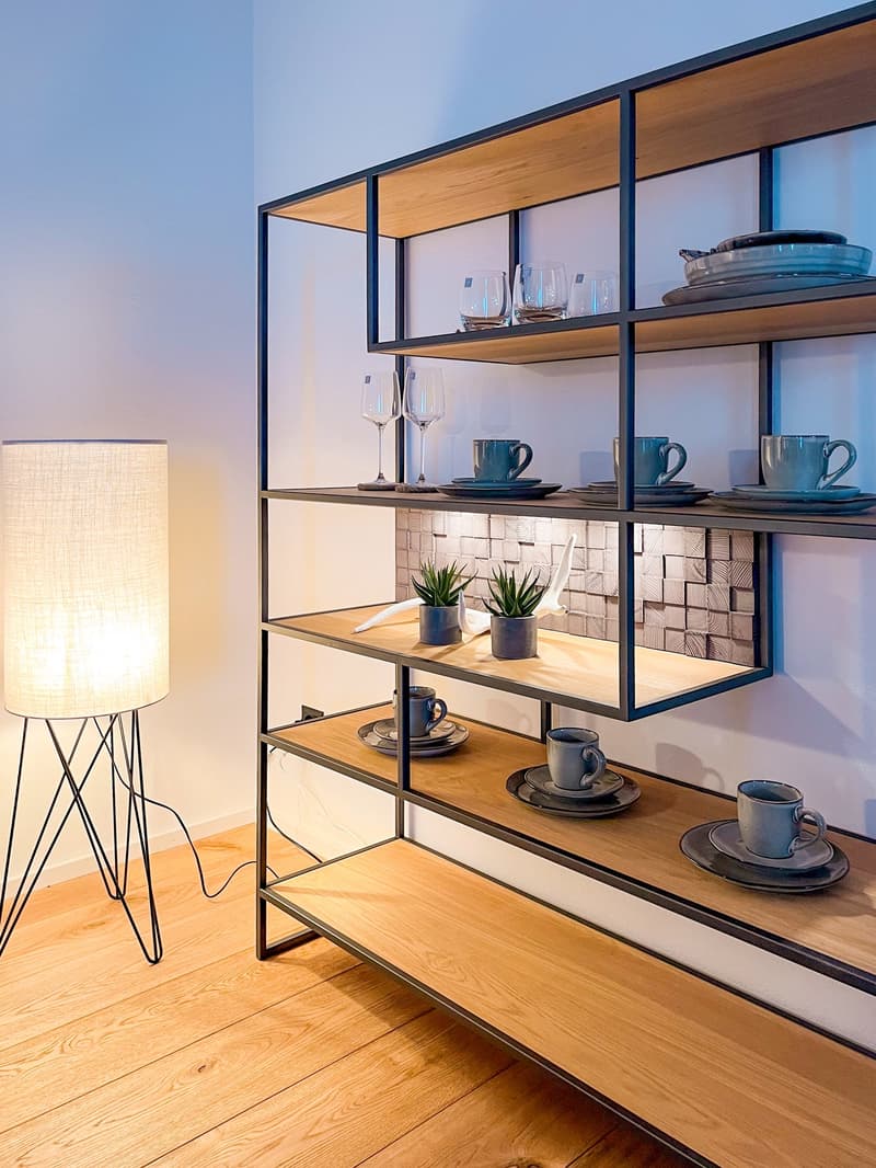 A shelf unit in a room with various plates, cups, wine glasses, and small potted plants. A floor lamp on the left side of the shelf and an electrical outlet at the bottom of the shelf.