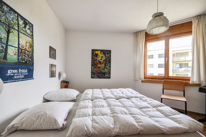 white walls, white bedspread, bed, 2 pillows, lamp, 3 paintings, chair, window, piano