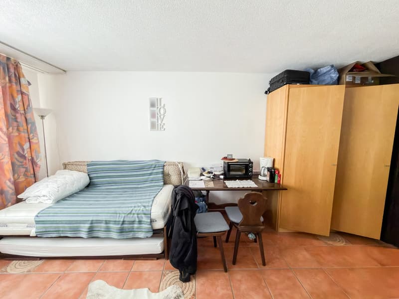 small room with a bed, desk, two chairs, cabinet with books, microwave, and storage boxes