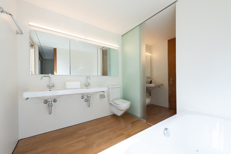 white modern bathroom with white tiles, two sinks, toilet, walk-in shower, bathtub, wooden floor