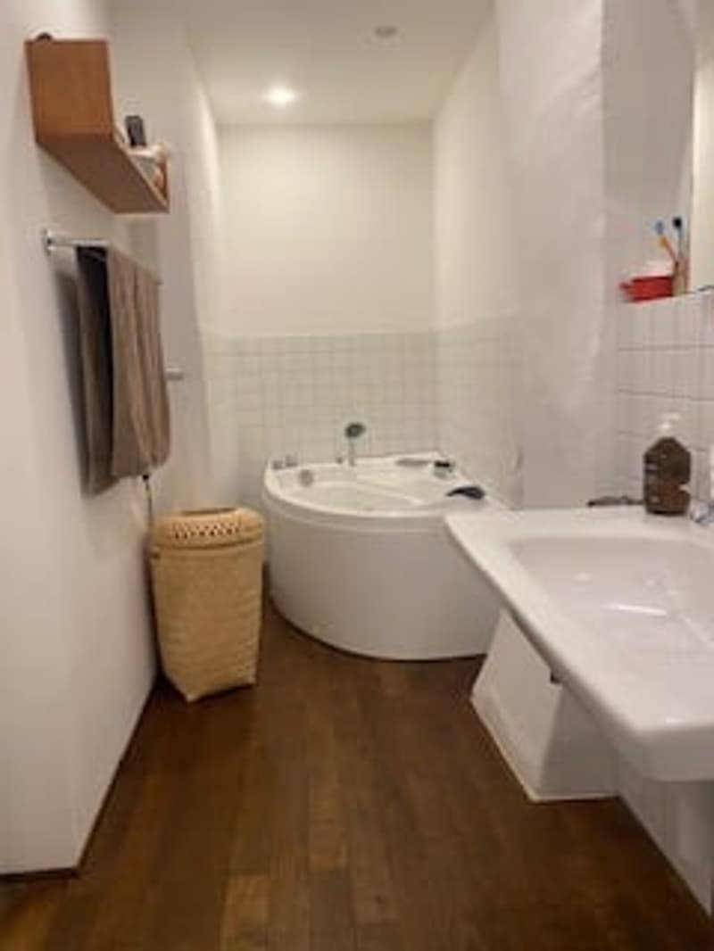 modern bathroom with white bathtub, wooden floor, white walls, white sink, wooden towel basket, shower head