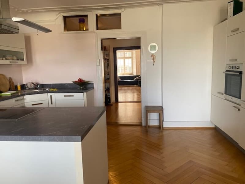 Modern open-plan kitchen with wooden floors, white cabinetry, central island, white walls, and ceiling, a large window, and an oven in the corner.