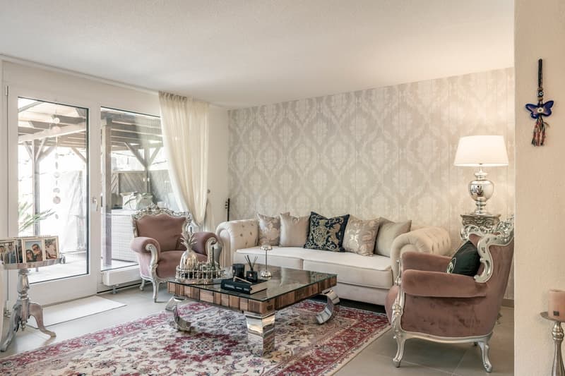 Spacious living room with large windows, patterned wallpaper, white sofa, pink armchair, glass coffee table, and decorative accessories.