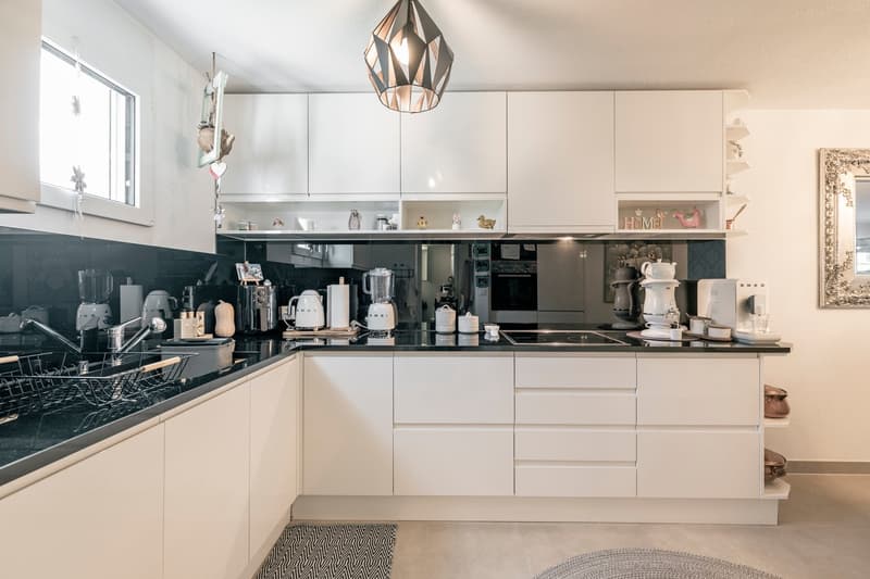 Spacious modern kitchen with white cabinets, black countertops, and various kitchen appliances and utensils. The kitchen features a dishwasher and a decorative pendant light fixture.