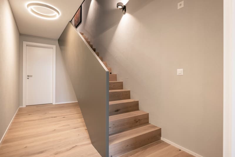 The image shows a hallway with a wooden staircase leading up. The walls are painted in a light, neutral color and there is a white door visible. The floor is made of hardwood planks. The lighting in the hallway is provided by a circular light fixture on t